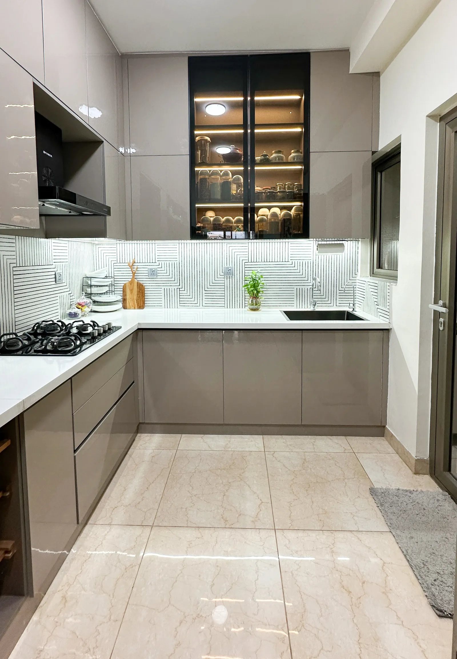 Taupe modular kitchen with patterned backsplash and glass display unit — Somani's Residence